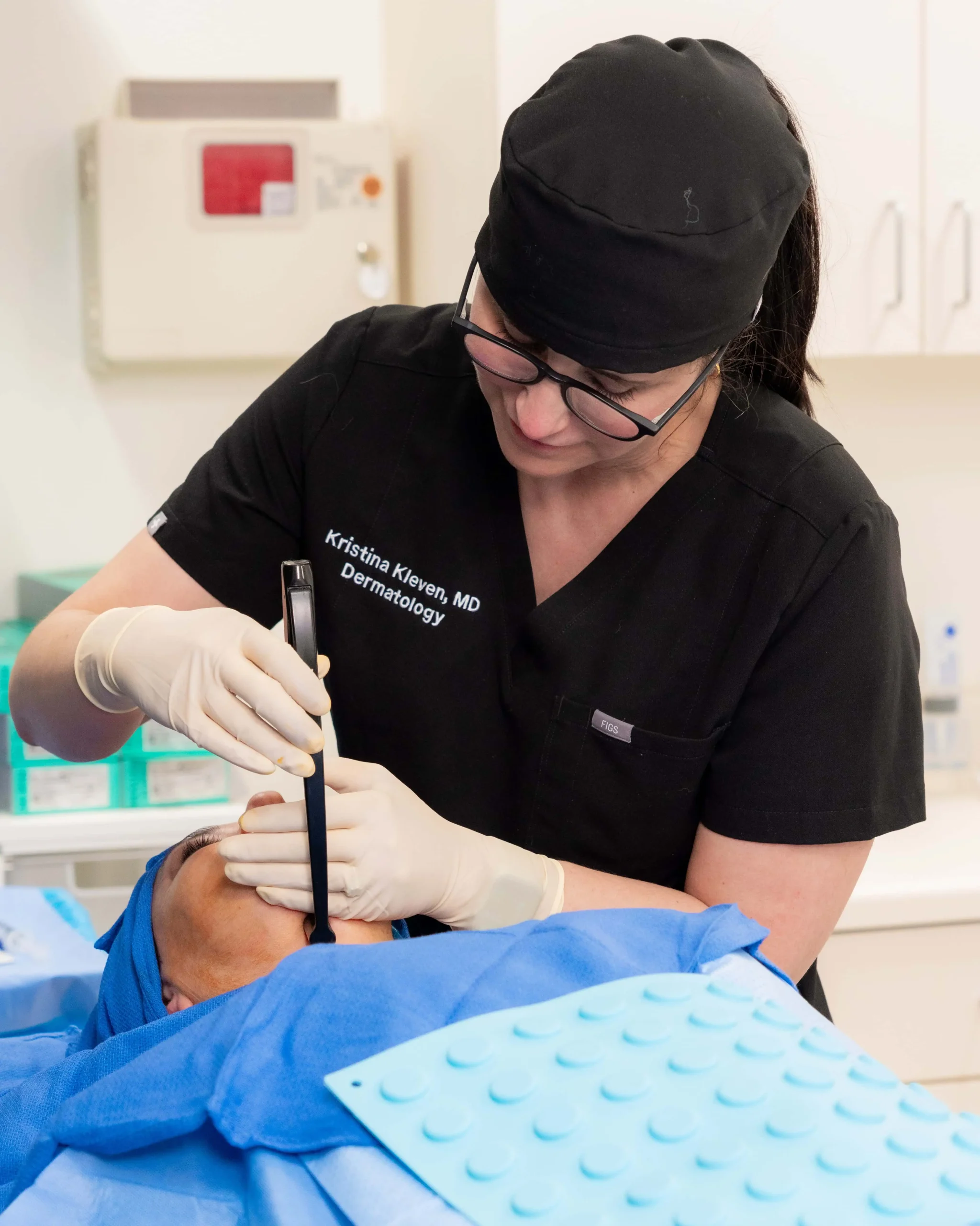 A dermatologist wearing black scrubs, a surgical cap, and gloves performing a facial procedure on a patient using a handheld medical device.
