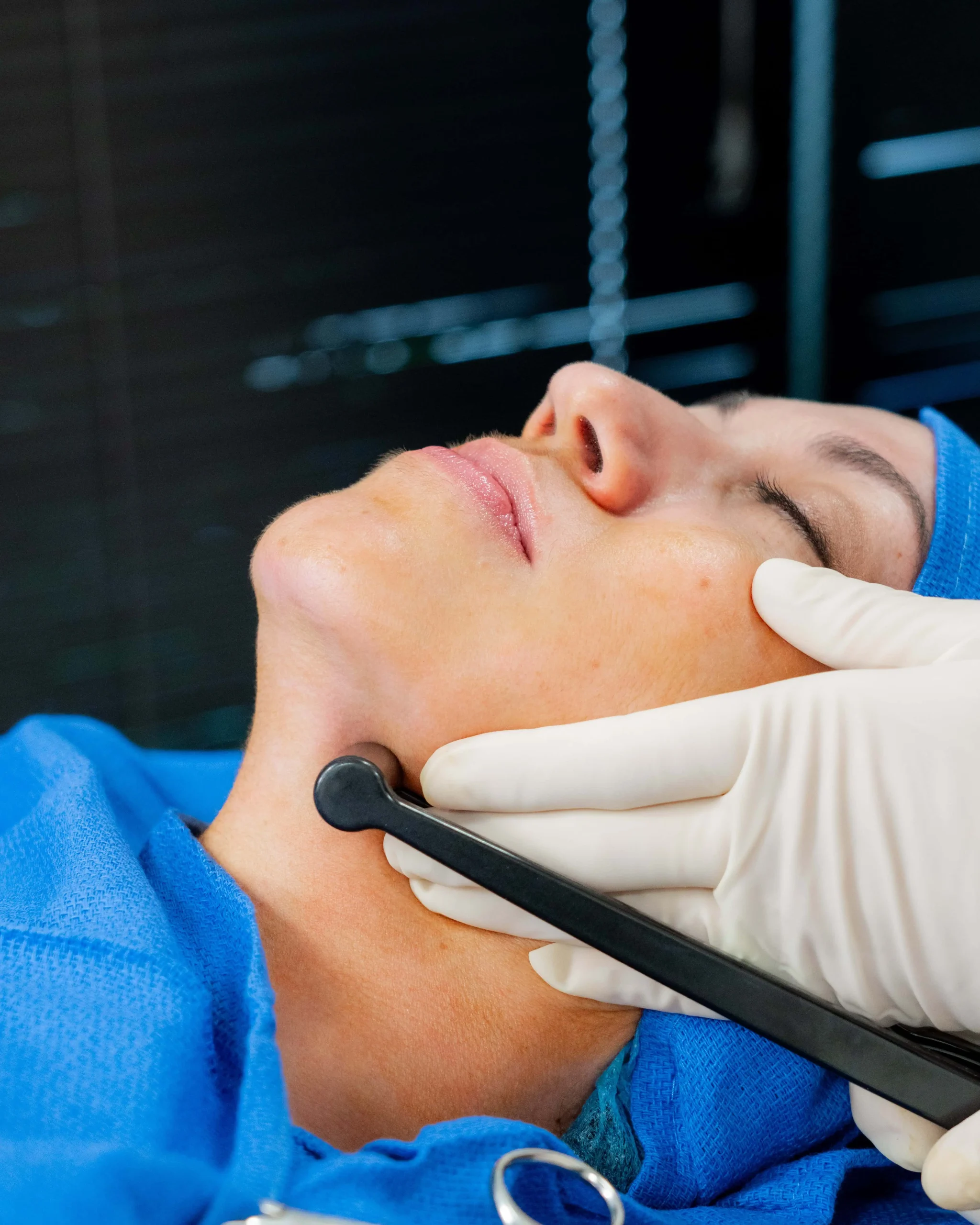 A close-up of a gloved hand using a black handheld medical device on the neck of a reclining patient wearing blue surgical attire.